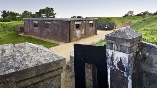 The Tool Store seen from the entrance gates at Reigate Fort, Surrey. A short square building surrounded by grass, with gates in the foreground.